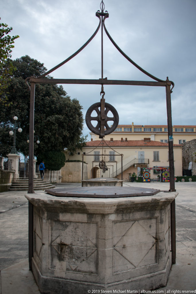 Stone wellhead in a paved square with a tall iron frame and suspended pulley wheel above it.