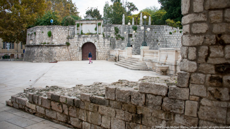 Paved square with low ruins, curved stone bastion wall, round-arched gate, balustrade, and trees above.