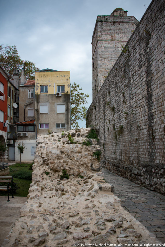 Low archaeological stone foundations beside a tall city wall and square defensive tower, with apartment buildings beyond.