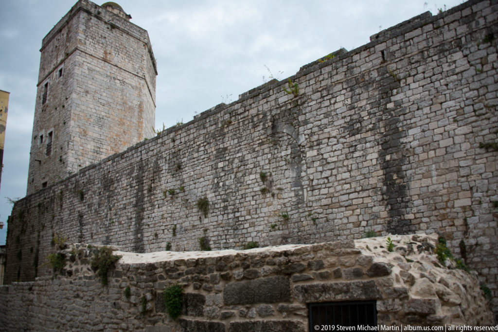 Massive limestone city wall with tall square tower and lower archaeological masonry in the foreground.