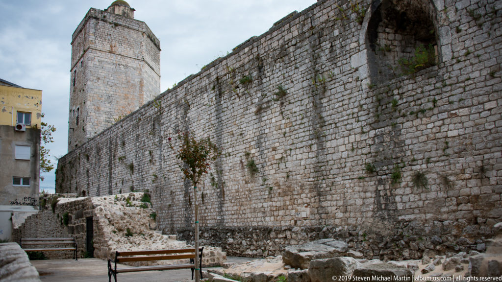 Long stone defensive wall with tall square tower and exposed archaeological masonry at its base.