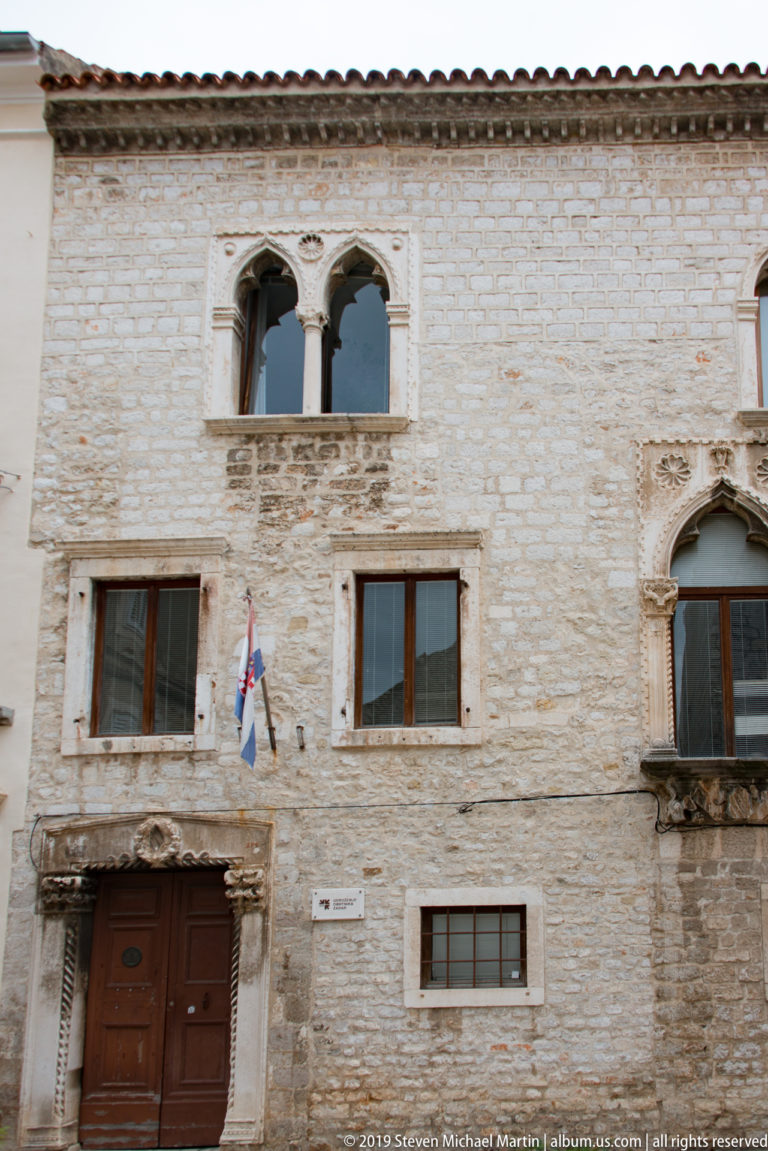 Historic stone façade with carved doorway, paired Gothic upper window, Croatian flag, and irregular rectangular openings.