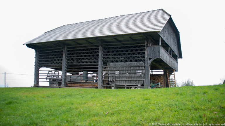 Open-sided wooden hay barn with slatted walls, broad pitched roof, and stored agricultural material on a grassy hill.