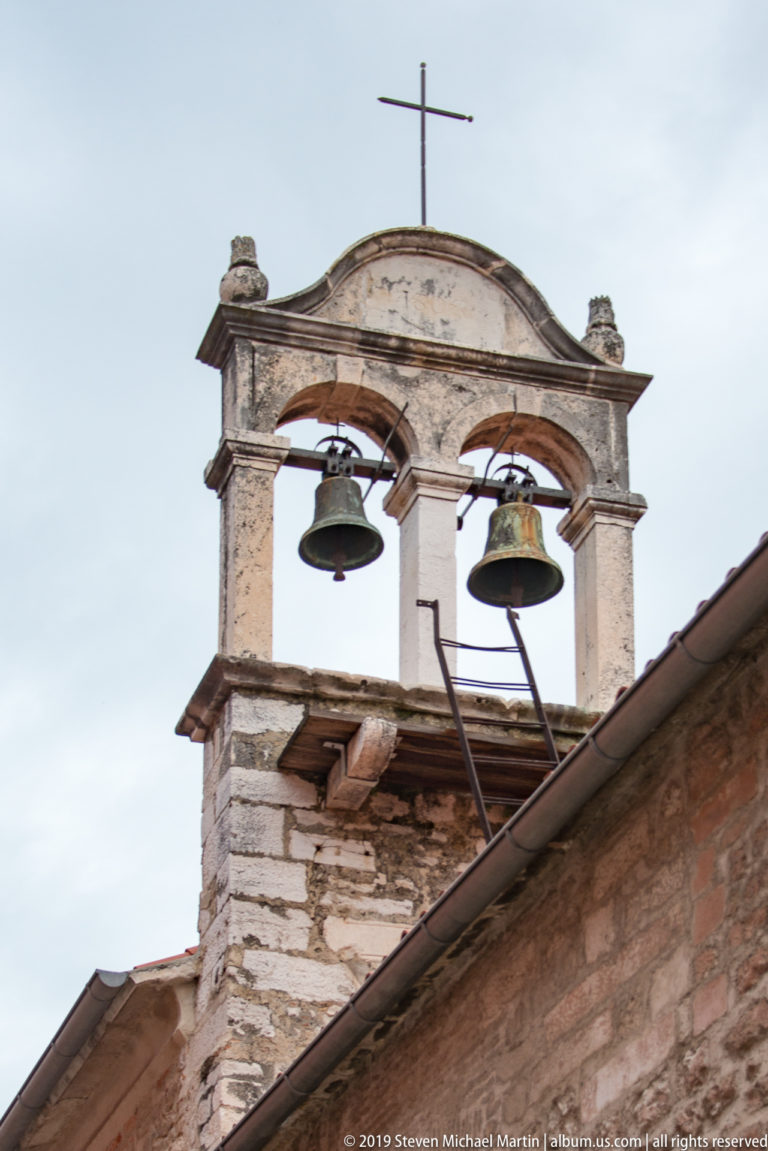 Two bronze bells hanging in a weathered double-arched stone bellcote topped by a cross.