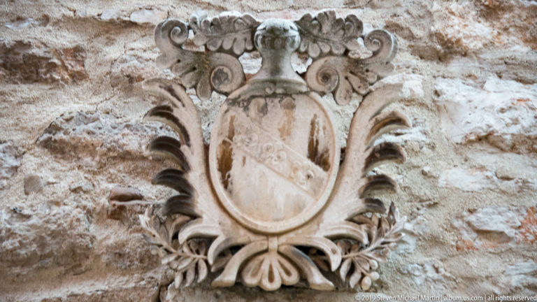 Ornate carved stone cartouche with oval shield, scrolling foliage, and a small sculpted head on rough masonry.
