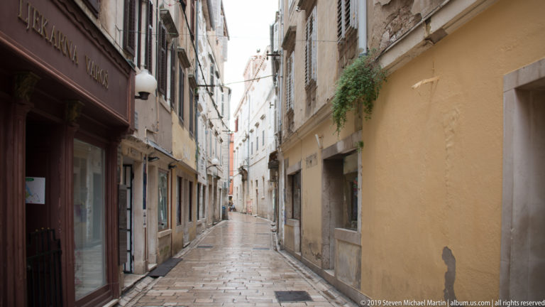 Narrow stone-paved pedestrian street lined with closely spaced historic buildings, shuttered windows, shops, and hanging signs.
