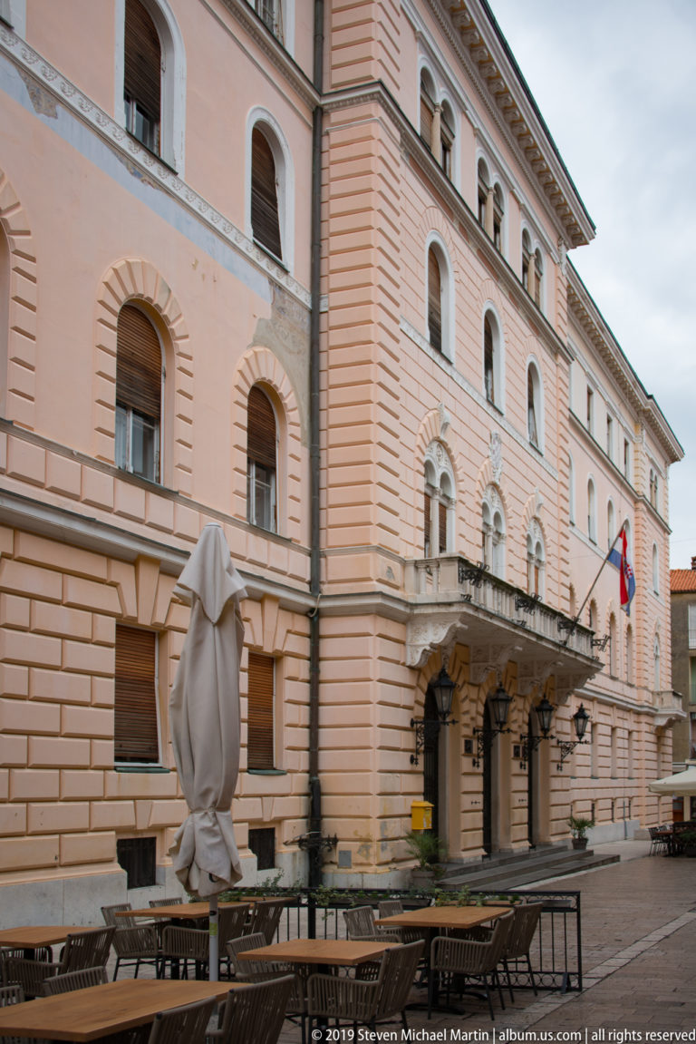 Long salmon-colored civic building with arched windows, balcony, Croatian flags, and café seating in front.