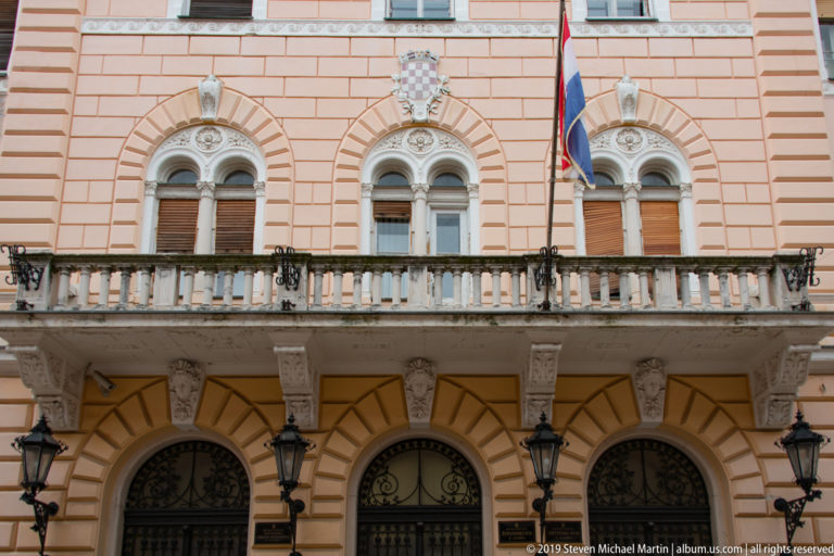 Upper civic façade with paired arched windows, balcony, Croatian coat of arms, and national flags.