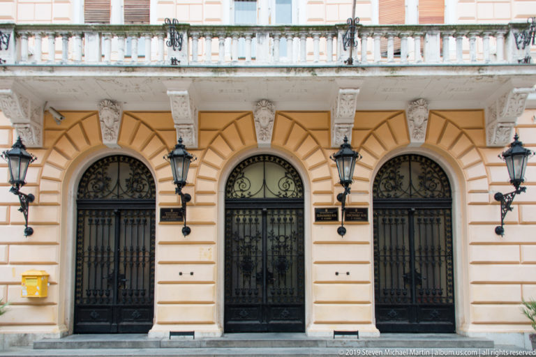 Three arched ironwork entrances beneath a balustraded balcony on an ornate salmon-and-cream civic façade.