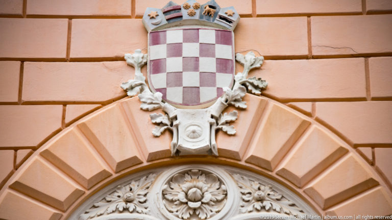 Croatian red-and-white checkerboard coat of arms with smaller crest shields above a rusticated stone arch.