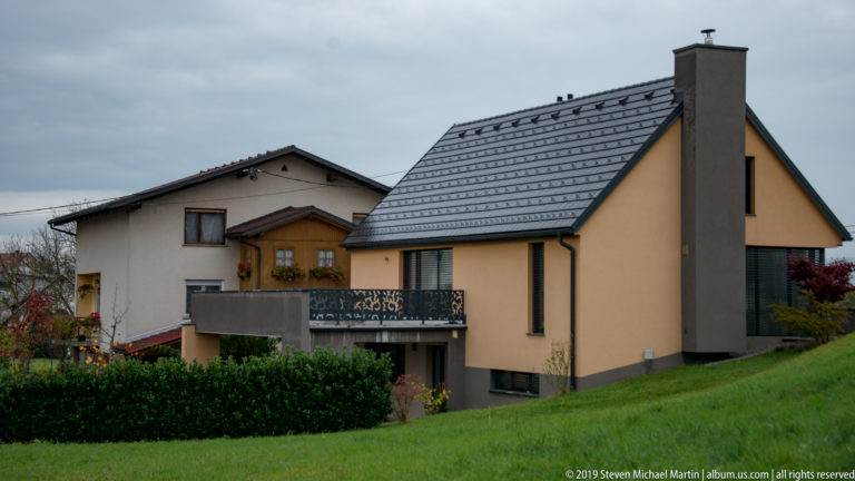 Modern detached house with steep gray roof, balcony, chimney, and neighboring home on a grassy slope.