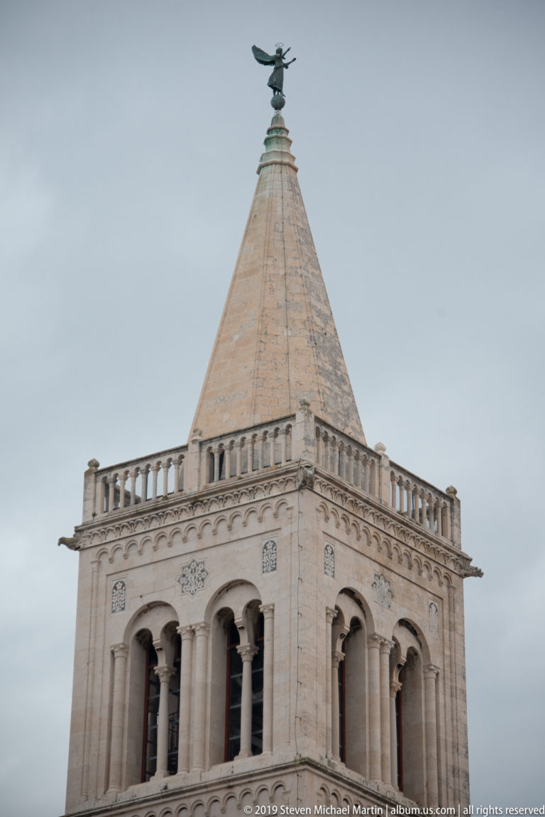 Upper arcaded stages and pyramidal roof of St. Anastasia Cathedral bell tower against a cloudy sky.