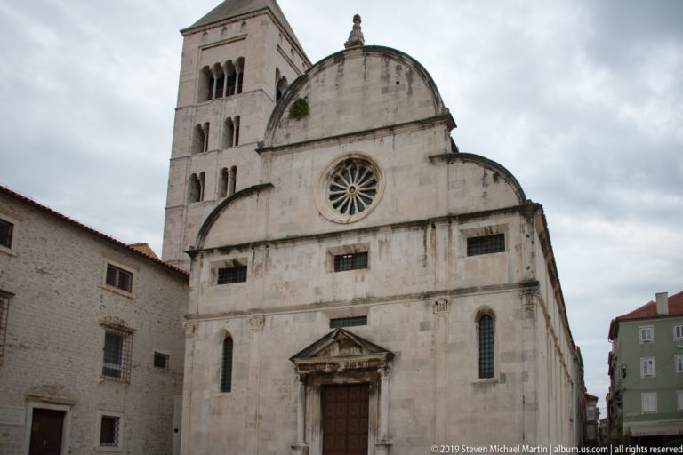 Stone church façade with central rose window, curved gable, dark doorway, and bell tower rising behind at left.