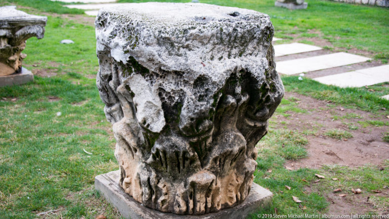 Weathered carved stone capital with leaf and volute ornament resting on grass among archaeological remains.