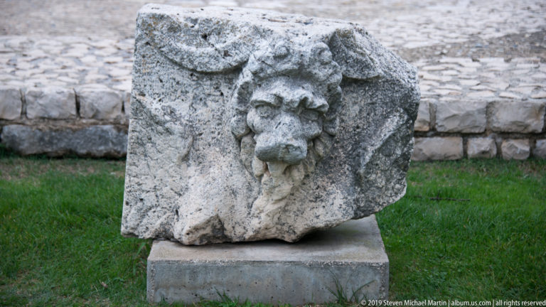 Weathered stone block with a projecting carved lion or feline head and traces of surrounding ornament.