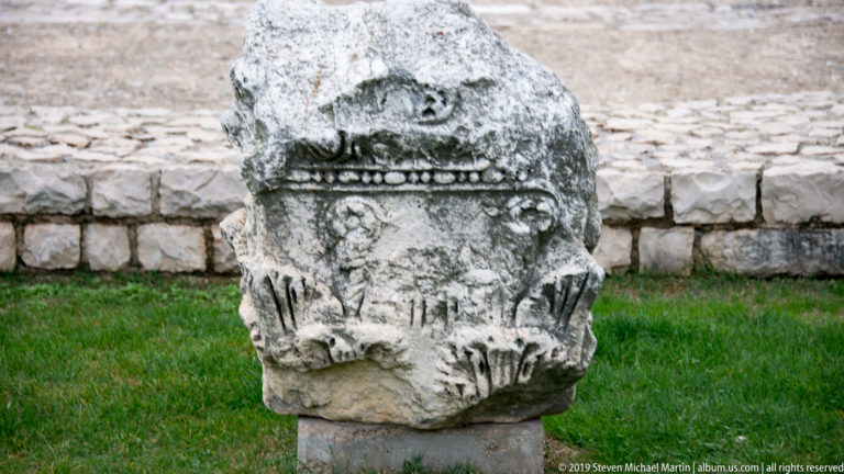 Weathered Roman stone fragment carved with a central face or mask, moldings, and dense vegetal ornament.