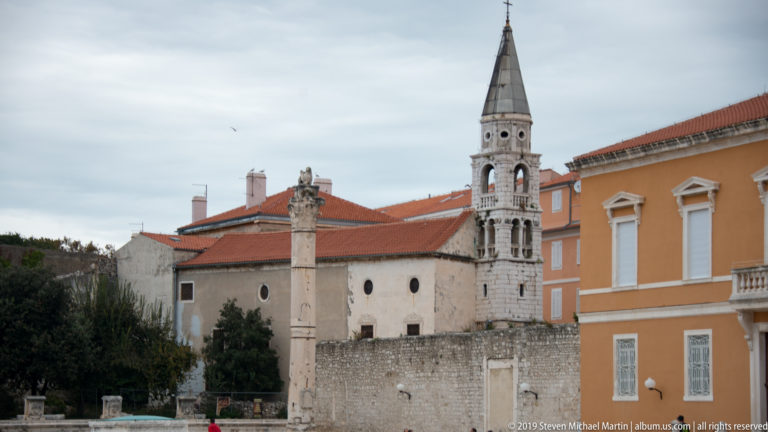 Roman column in Zadar’s forum with the arcaded bell tower of St. Mary’s Church and historic buildings behind.