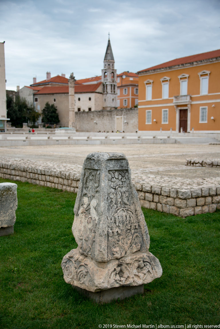 Large weathered Roman stone fragment with flared sides and carved vegetal ornament resting on grass in the forum.
