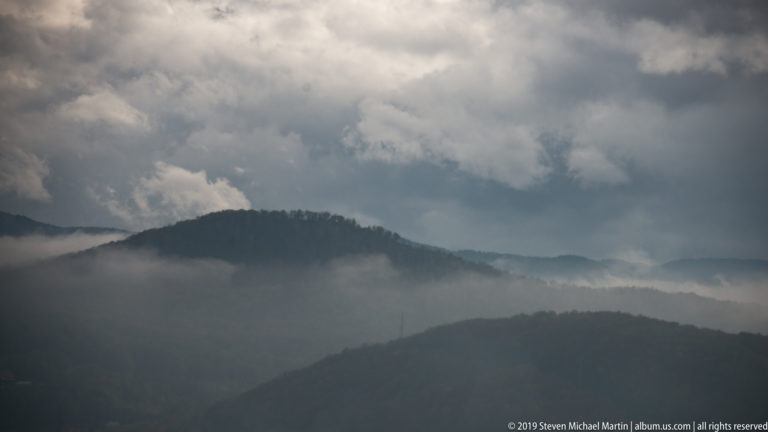 Dark forested ridge emerging through horizontal mist beneath heavy clouds, with additional hills fading into the distance.