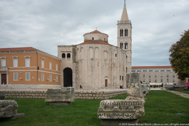 Church of St. Donatus seen across Roman Forum fragments with St. Anastasia Cathedral bell tower rising behind.