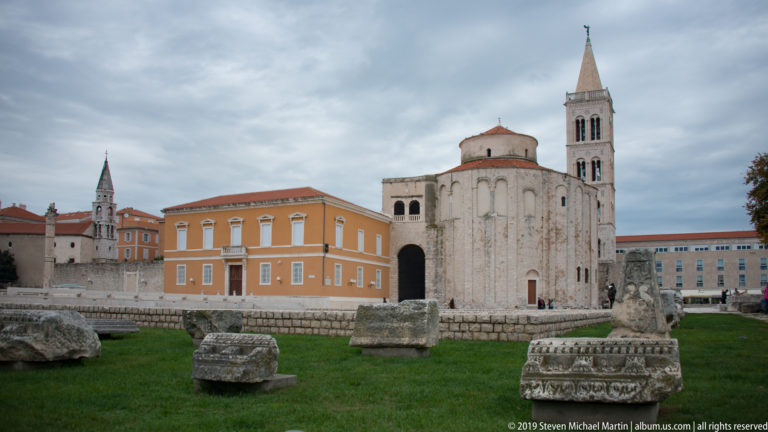 Rounded rear exterior of St. Donatus beyond Roman stone fragments, with a tall bell tower and orange civic building nearby.