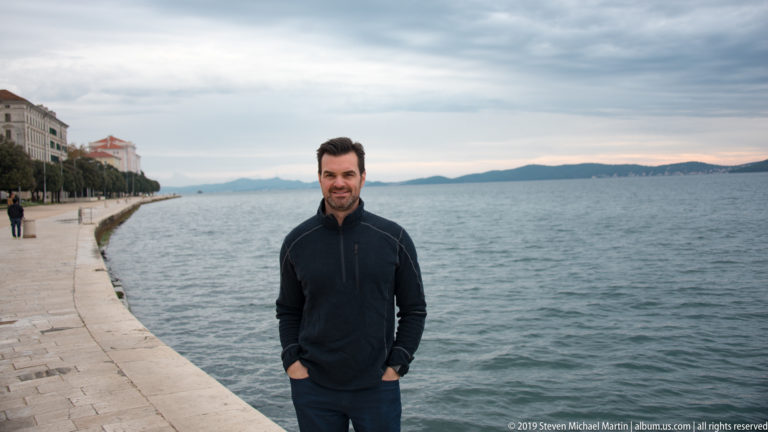 Man in a dark jacket standing on Zadar’s stone waterfront promenade with the Adriatic Sea and distant islands behind.