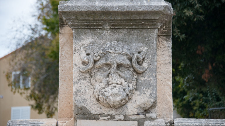 Ancient stone block carved with a weathered mask framed by curling horn- or volute-like forms.