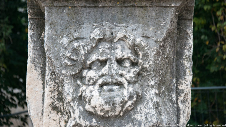 Close view of a weathered carved stone face with curling hair on an ancient architectural block.