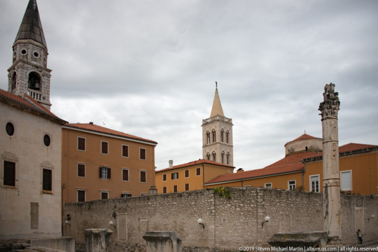 Roman Forum ruins in Zadar with scattered stone fragments, a tall ancient column, church towers, and red-tiled roofs behind.