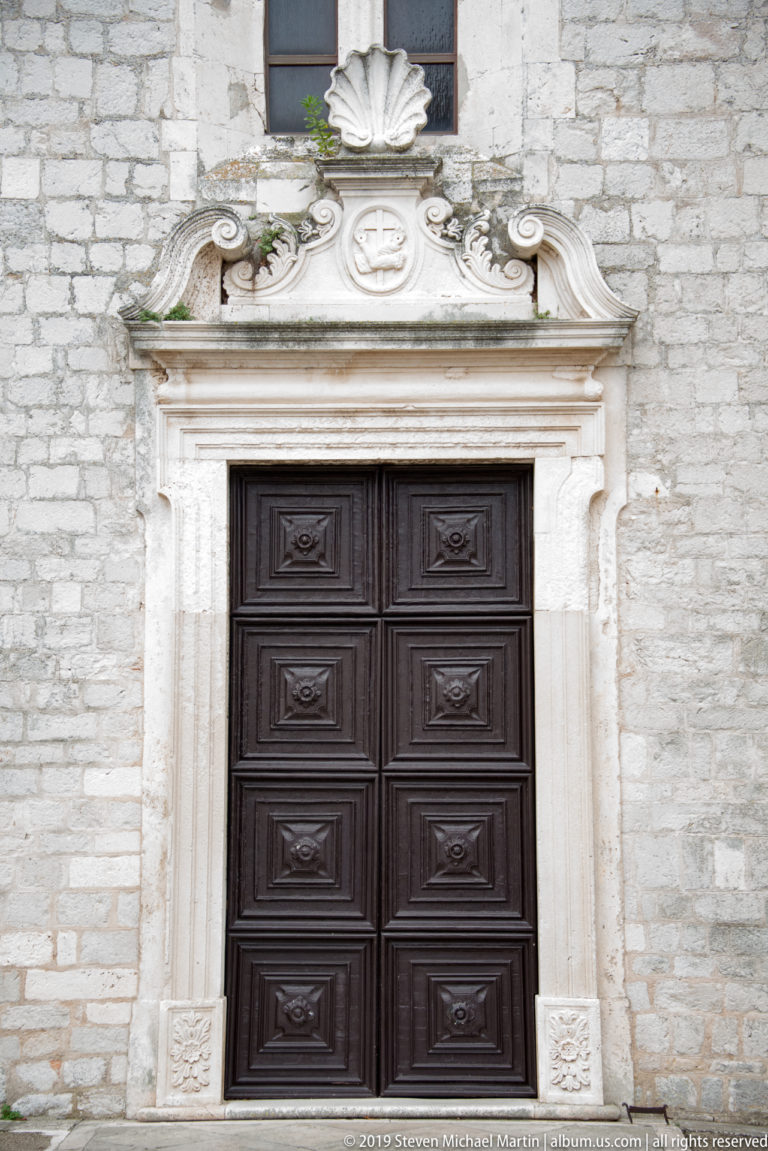 Dark wooden double door in an ornate pale-stone portal topped by scrollwork, roundel, and shell motif.