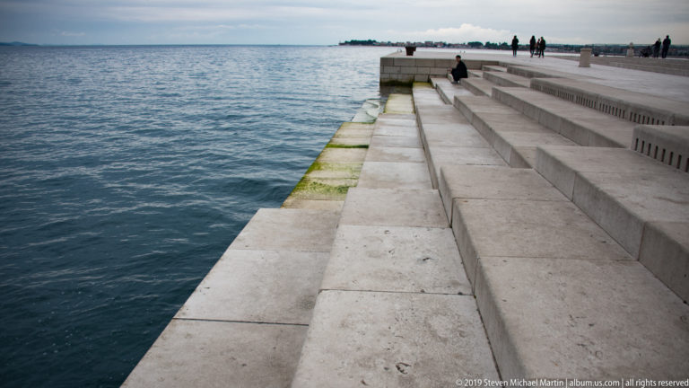 Wide limestone steps of Zadar’s Sea Organ descending beside the Adriatic, with visitors along the upper promenade.