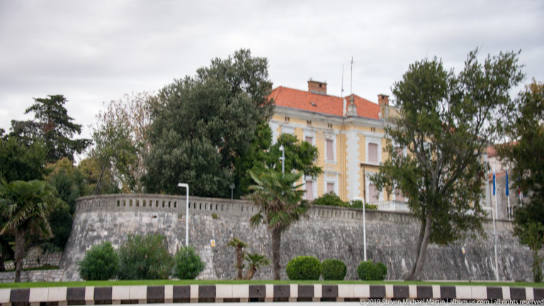 High stone fortification wall topped by landscaped trees and palms, with a pale historic building behind.