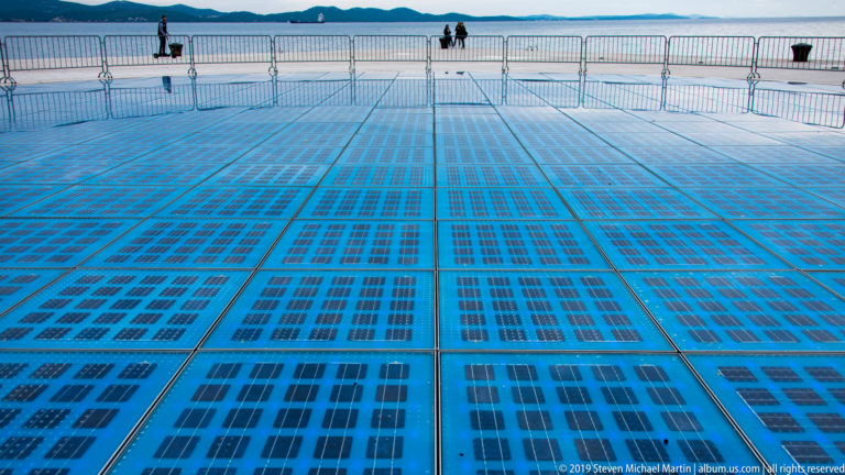 Close low-angle view across blue photovoltaic glass panels of Zadar’s Greeting to the Sun beside the sea.