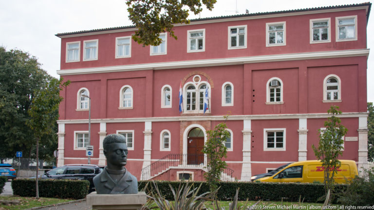 Red three-story institutional building with arched windows, central stairs, flags, and a portrait bust in front.