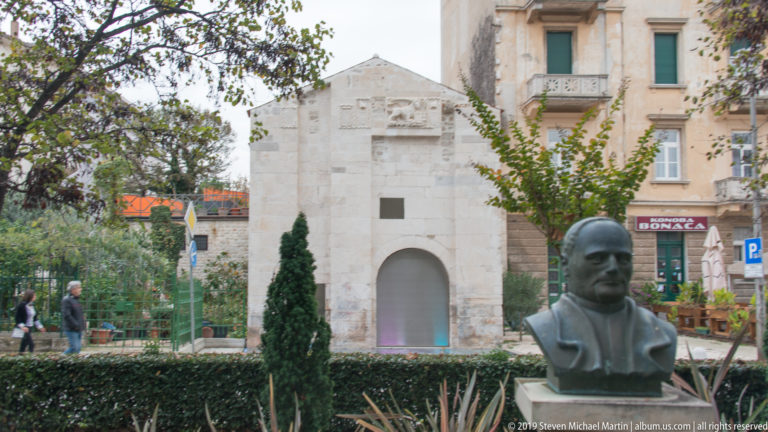 Bronze male portrait bust in a garden before a small pale-stone structure with a round-arched opening.