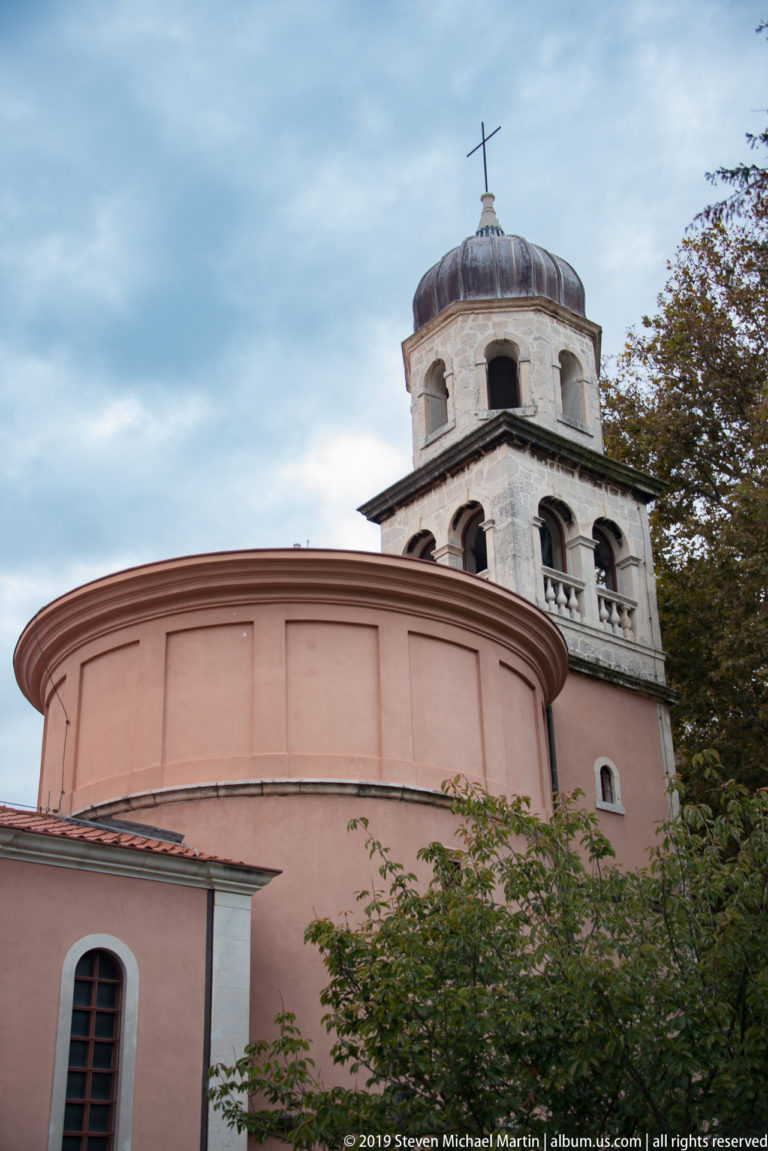 Large salmon-colored semicircular church apse with pale stone bell tower, domed roof, and cross behind.