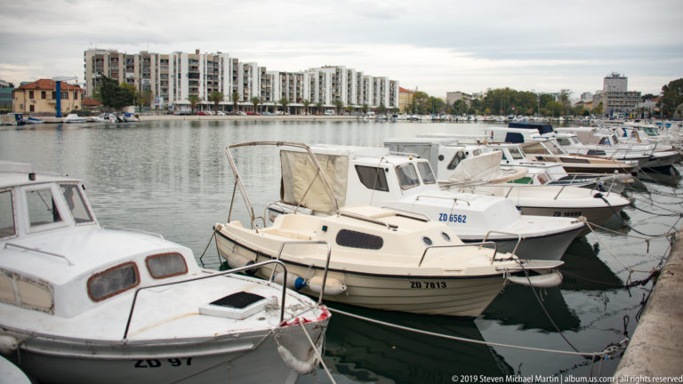 Rows of small cabin boats and motorboats moored in a calm Zadar harbor basin with modern waterfront buildings beyond.
