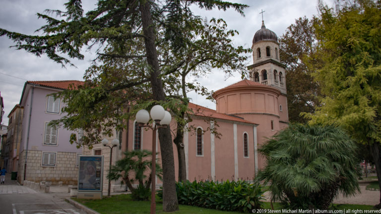 Pink rounded church apse with narrow arched windows and a square stone bell tower rising behind trees.