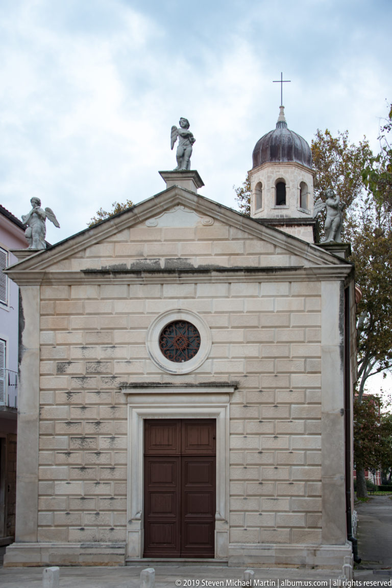 Small pale-stone church façade with pediment sculptures, oval window, dark wooden doorway, and domed lantern behind.