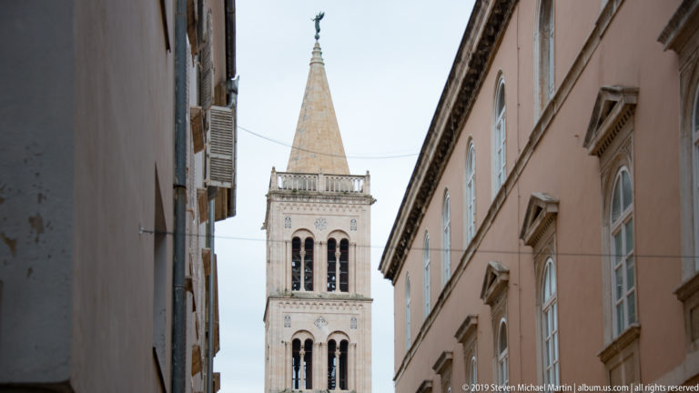 St. Anastasia Cathedral bell tower framed between buildings, with open arcades and a steep pyramidal spire.