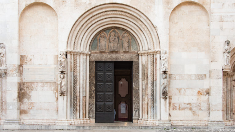 Main portal of St. Anastasia Cathedral with concentric carved arches, colonnettes, sculpted lunette, dark doors, and flanking figures.