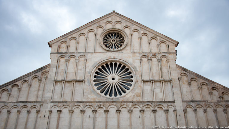Upper stone façade of St. Anastasia Cathedral with large lower rose window, smaller upper rose, blind arcades, and triangular gable.