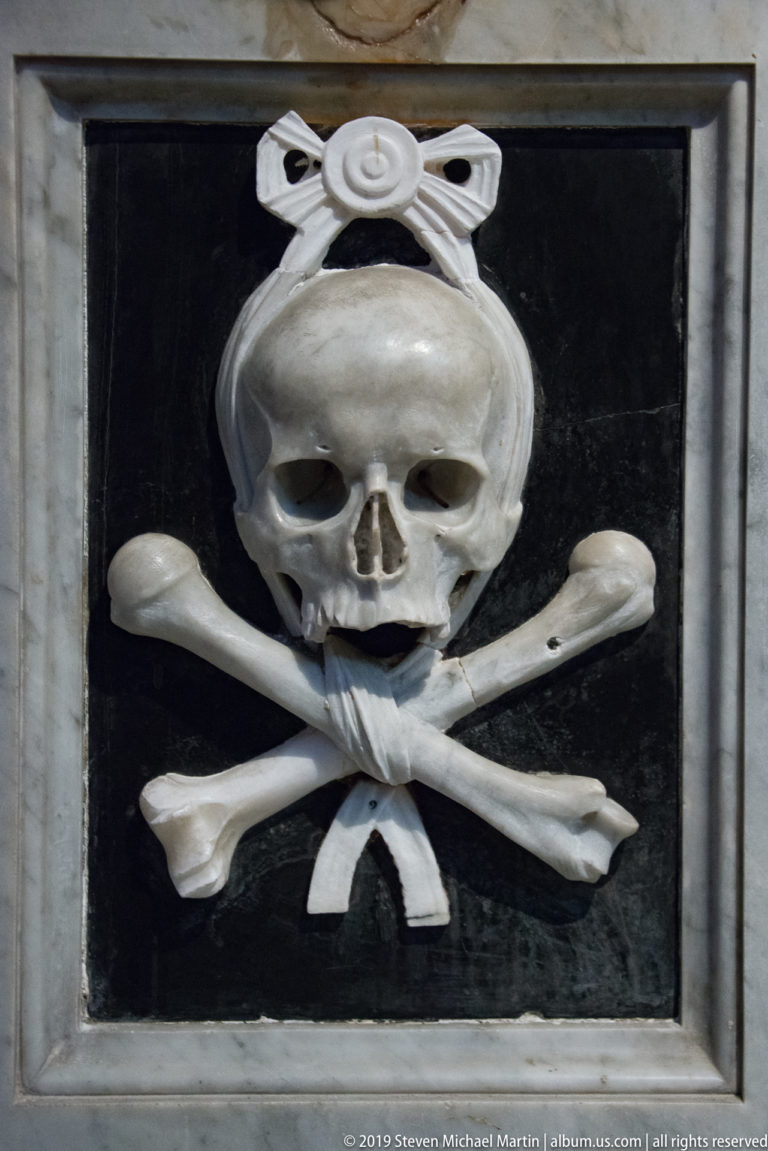 Stone skull and crossed bones on a dark panel with carved ribbon-like ornament above.