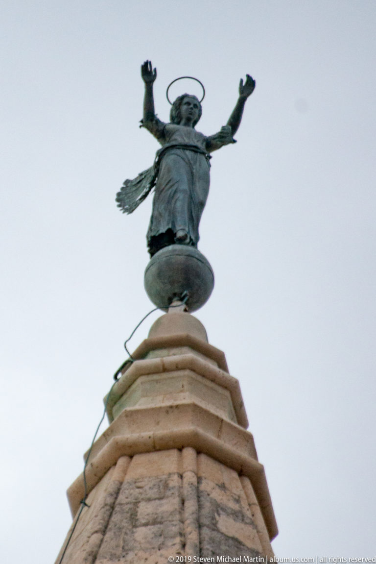 Bronze winged figure with raised arms and halo-like ring standing atop a pale stone pedestal.