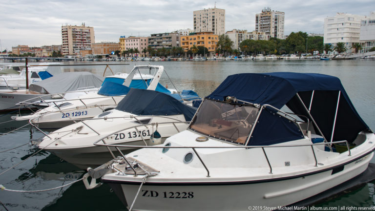 White recreational motorboats with canvas covers moored closely together in Zadar marina, with apartment blocks across the water.