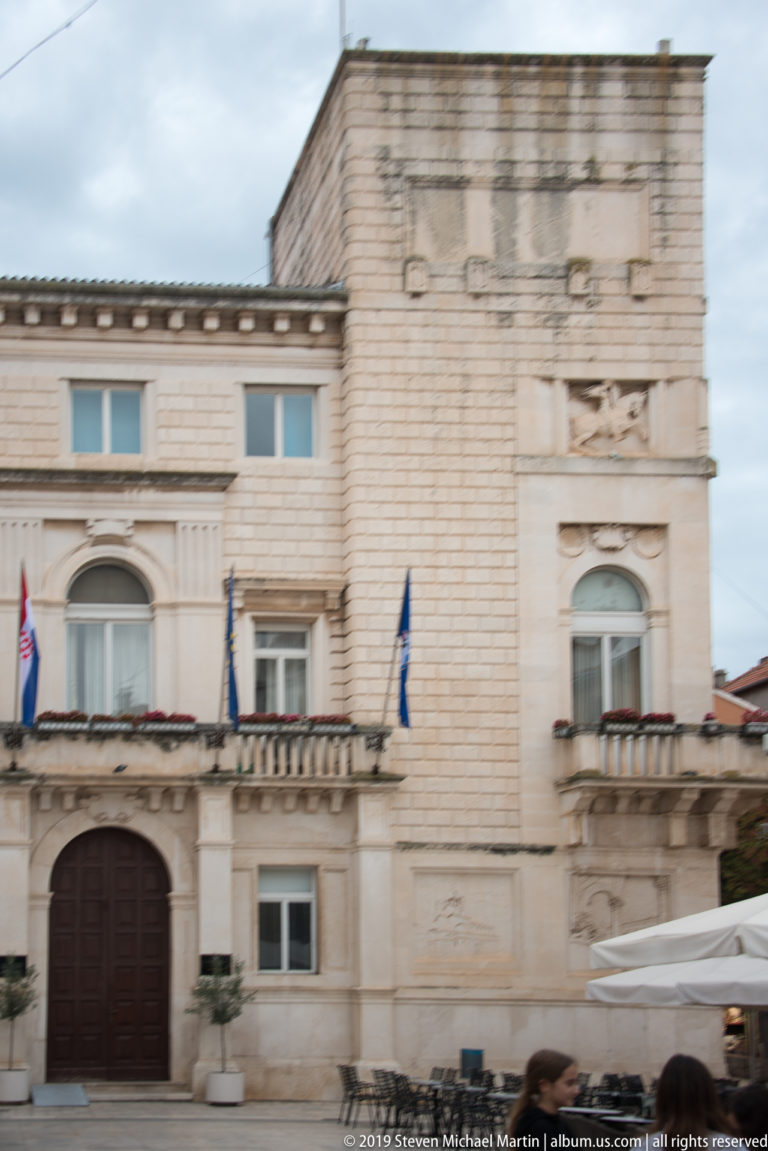 Stone civic building with central doorway, flags, rectangular windows, balustrade, and carved façade details.