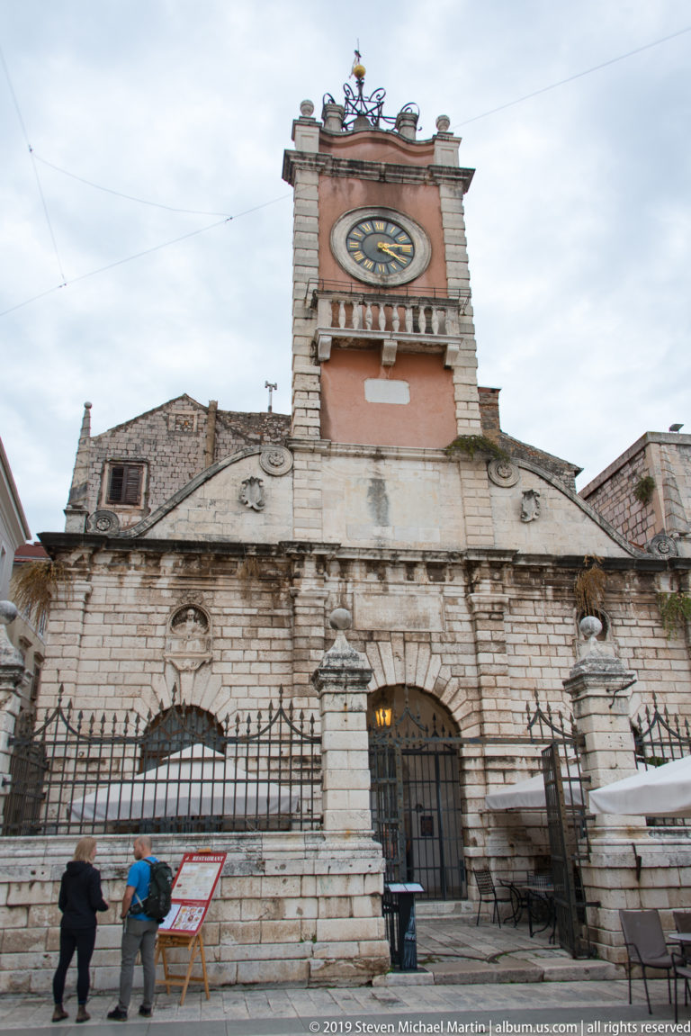 Frontal view of Zadar’s City Guard with stone guardhouse, arched openings, iron fence, and clock tower.