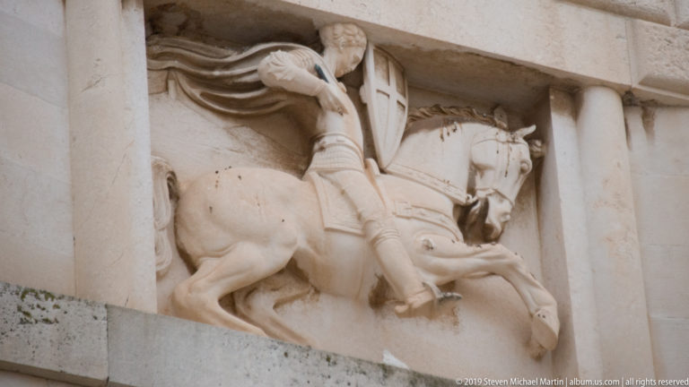 Carved stone relief of a rider on horseback beneath a flowing banner or drapery.
