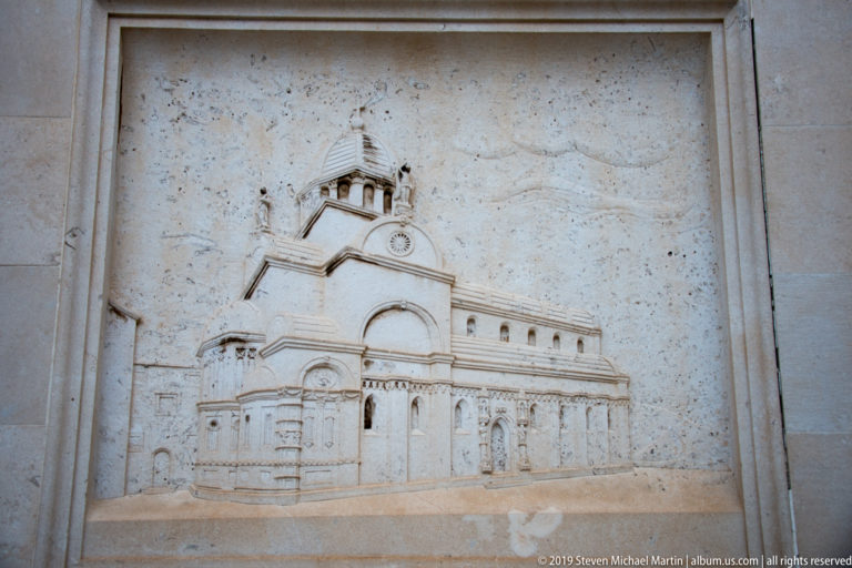 Weathered stone relief showing a church with central dome, arched façade, side volumes, and rows of windows.