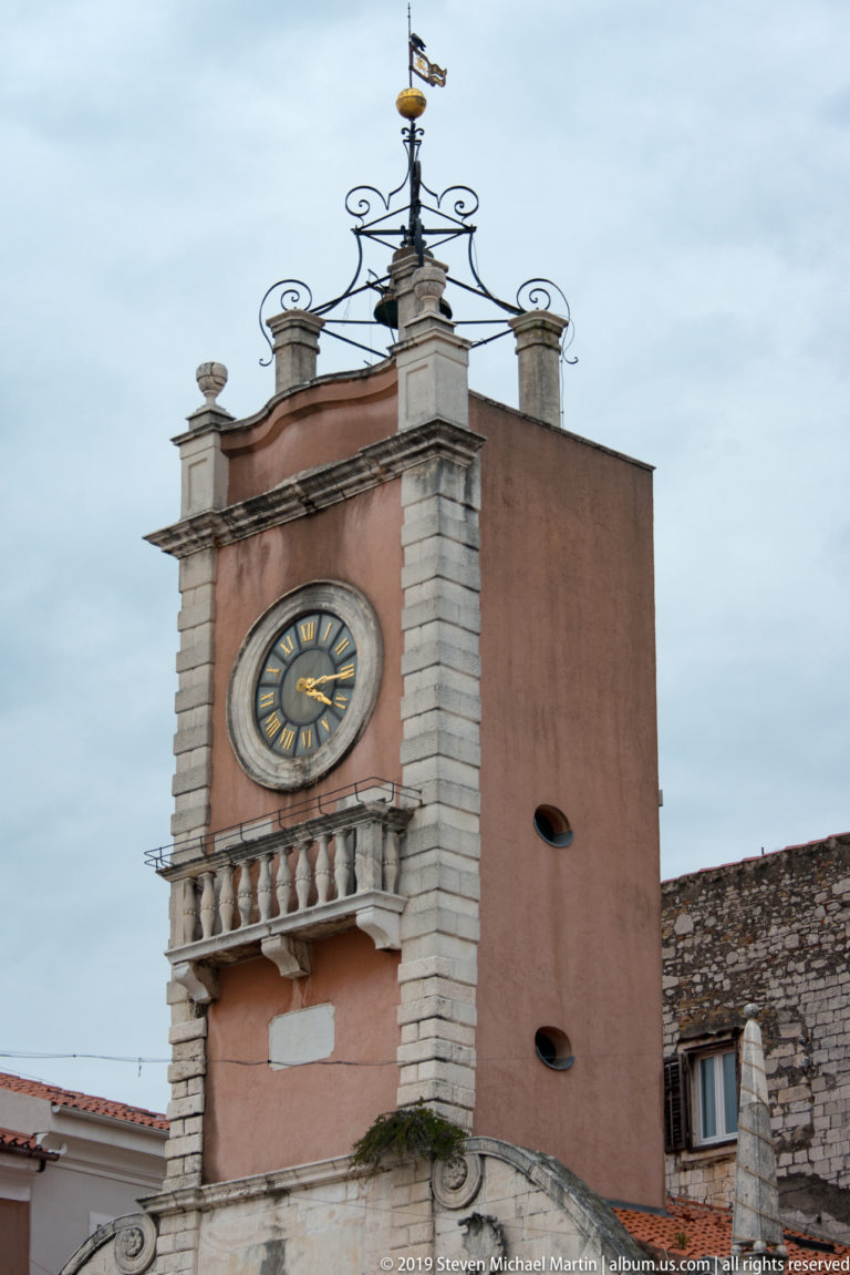 Salmon-colored clock tower of Zadar’s City Guard with round clock, stone trim, balustrade, and iron crown.